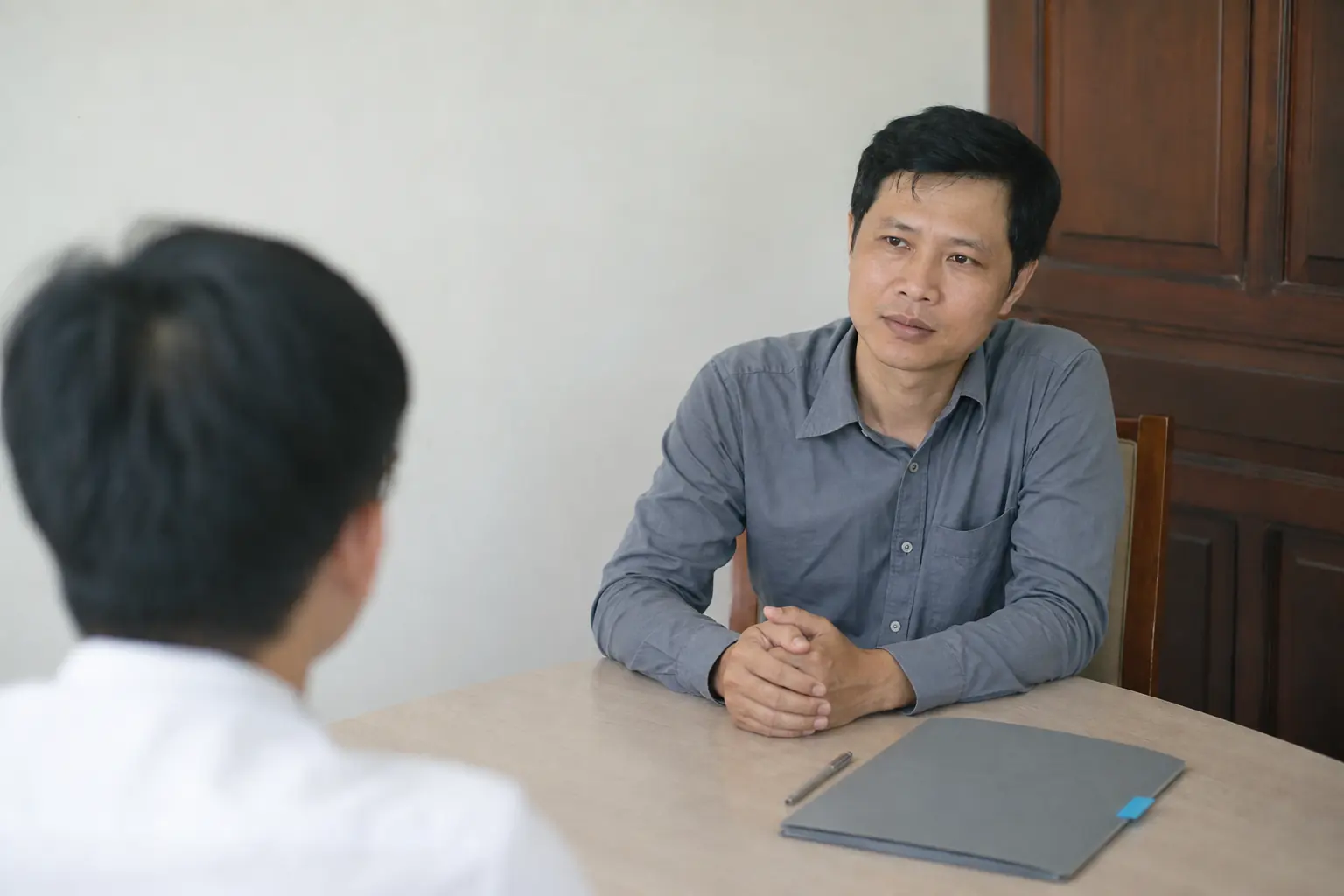 A quiet one to one interview across a plain table, a closed folder and a pen between the two people.